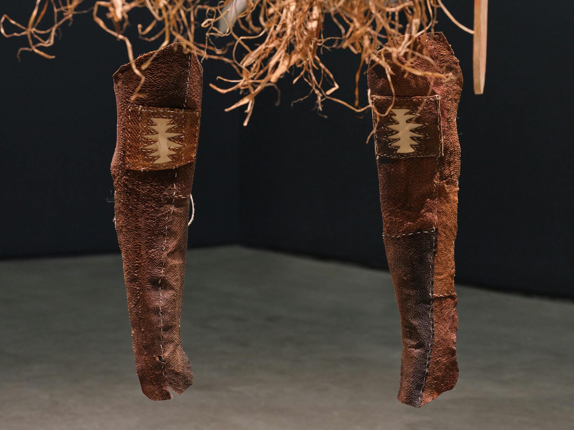 a pair of fishskin leather leg covers suspended behind the bottom of a cedar war apron. the knees of the leg covers feature a zig-zag patterned shape