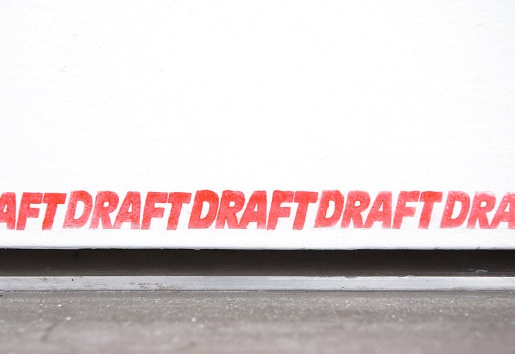 Detail of a work titled DRAFT by Ceal Floyer. A word, DRAFT has been repeatedly stamped onto the bottom of a white door, making a line with red ink.