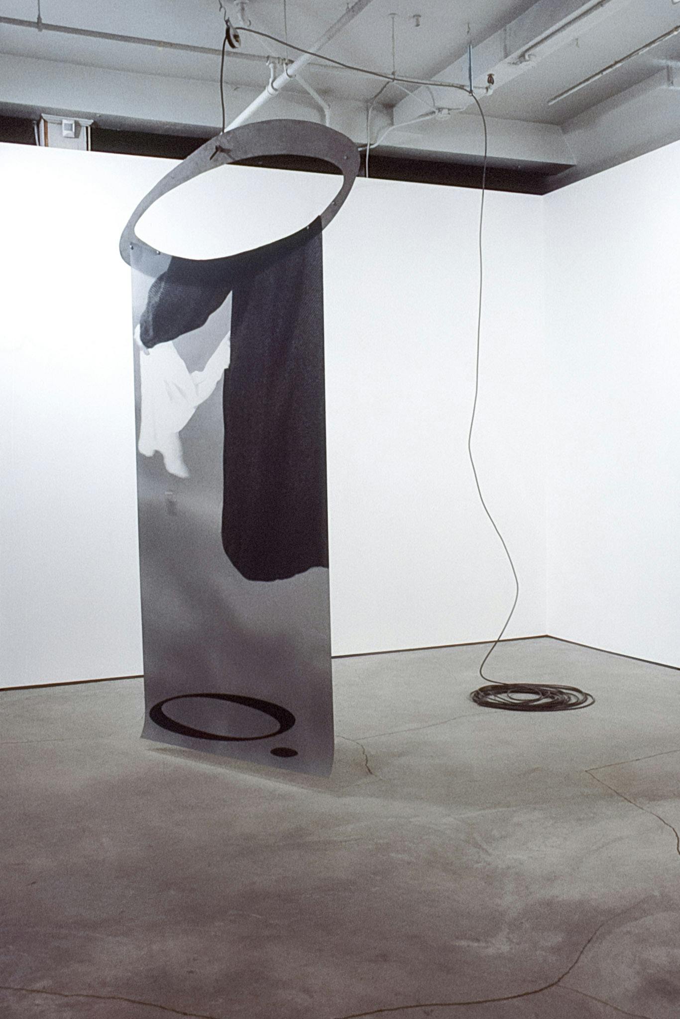 An artwork suspended in the corner of a gallery space. The work is composed of a photo printed on mylar, showing a person in a black robe, suspended from a cardboard oval hanging from a black cable. 