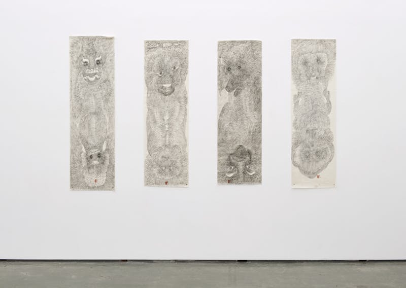 Four black and white drawings on long sheets of paper installed on a gallery wall. Each has faces resembling animals depicted at the top and bottom of column-like, organic forms of repeating lines.