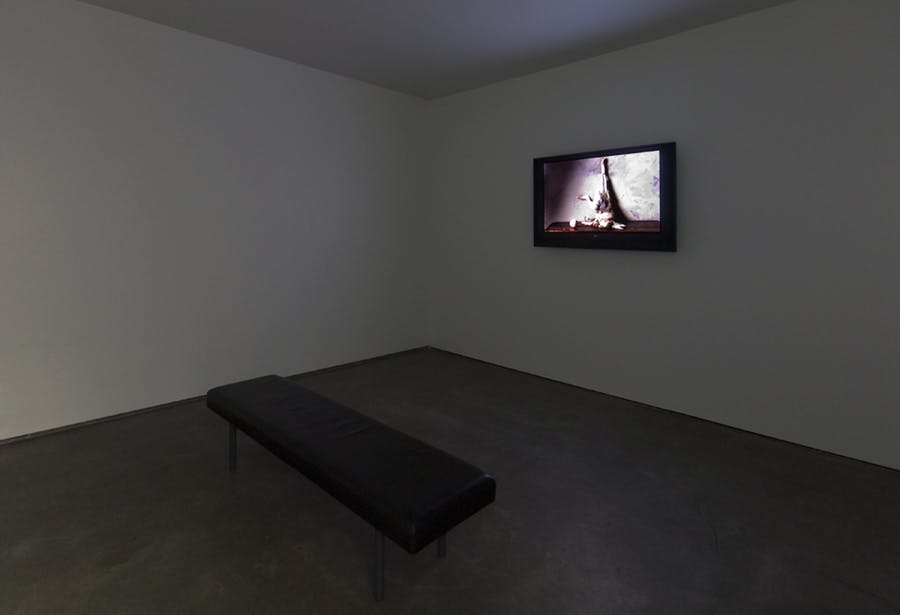 A monitor mounted on a wall in a dimly lit gallery displays an image. A black bench sits in the center of the room.