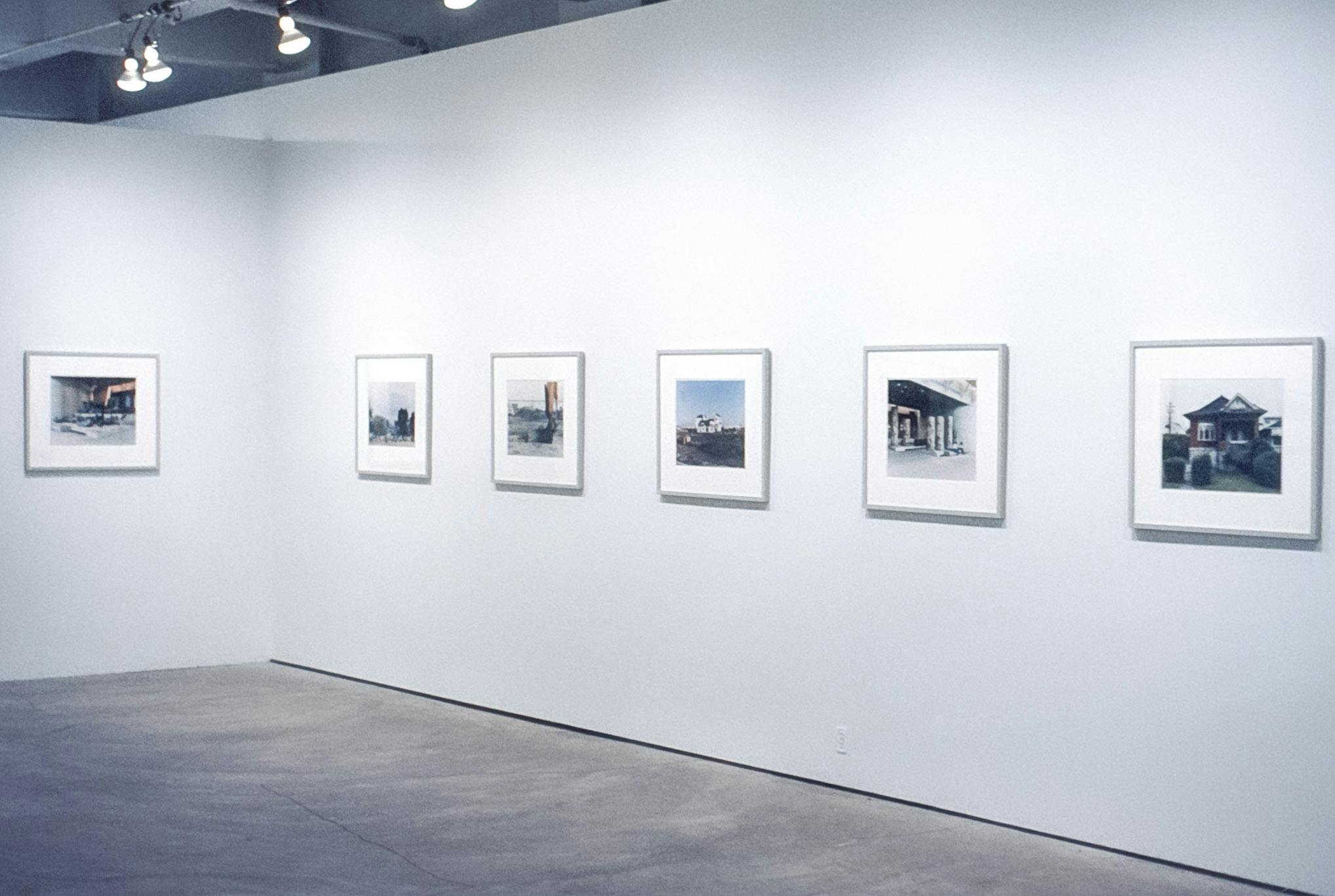 A gallery space with 6 framed photographic works mounted on the white walls. The works are both vertical and horizontal and depict city scenes, such as construction sites, houses, and underpasses.