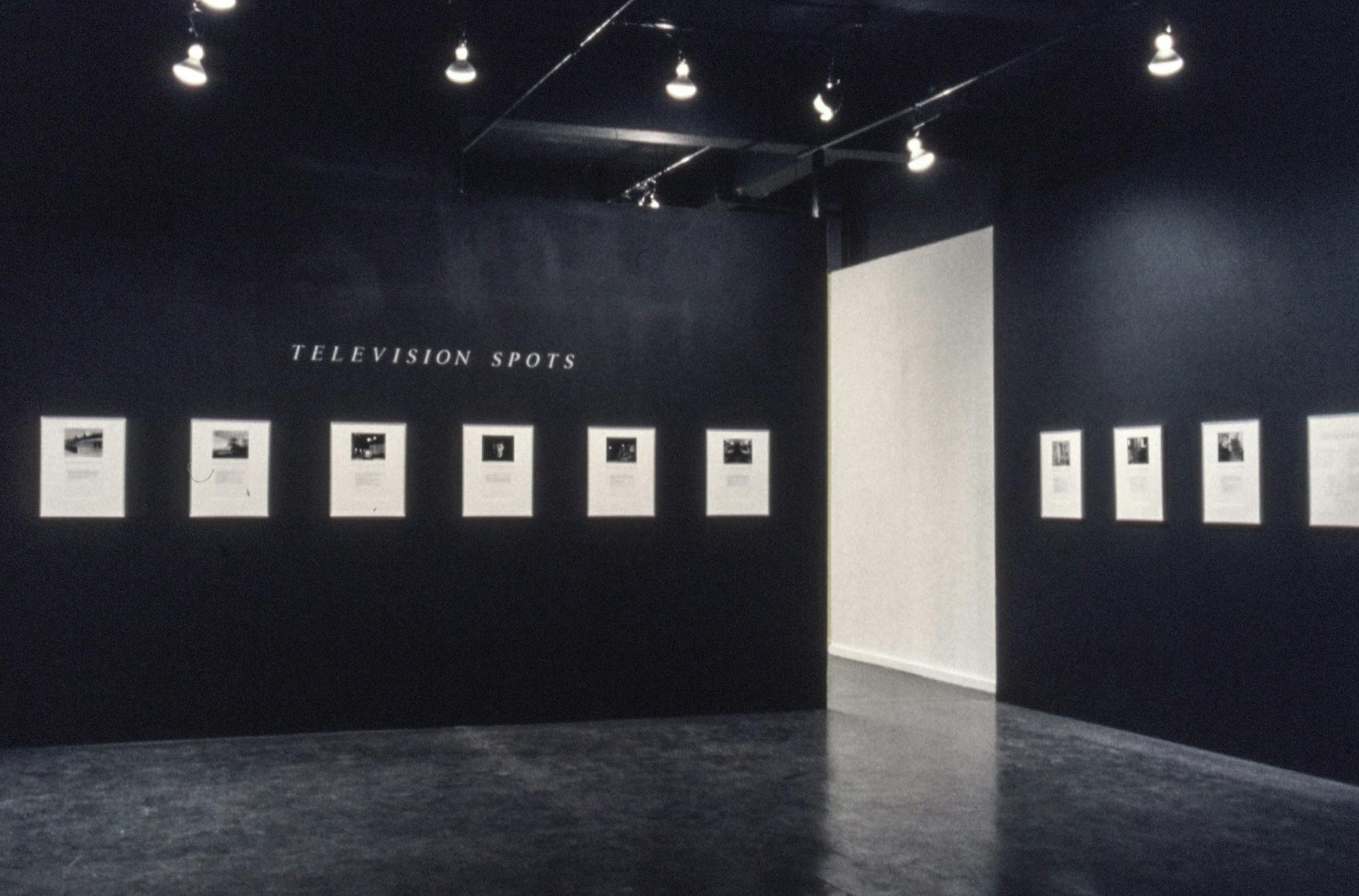 A gallery space with black walls. The wall shows large text reading "Television Spots" and several white artworks on the walls with black and white images and text. 