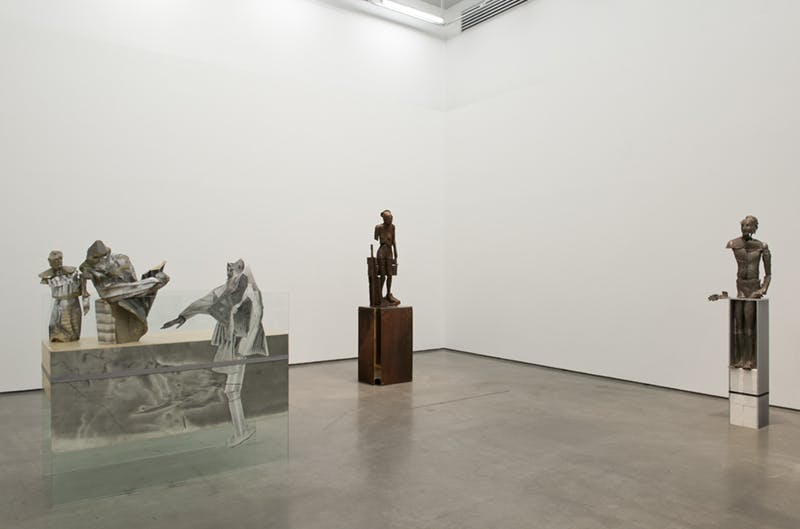 Five human form sculptures installed in a gallery on plinths made of boxes or a large stone slab with glass. Some of these sculptures have metallic surfaces while others appear to be made of stone.