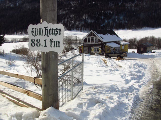 An image of a snowy landscape with a house made of log timber and forested hills behind. In the foreground of the image is a wooden post with a sign that reads “EDM house 88.1 fm.” 