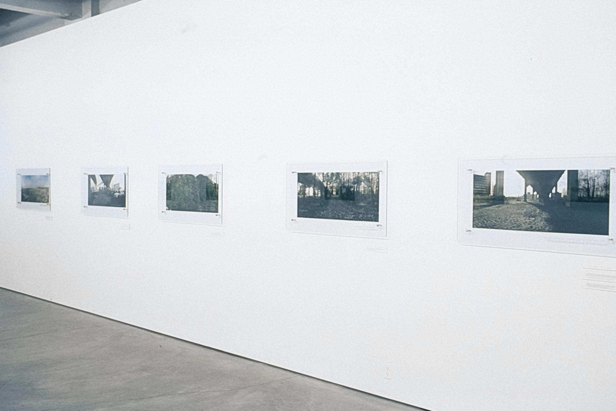 Several photos between sheets of glass mounted on a white wall. The photos show different natural and urban scenes around what is colonially known as the city of Vancouver.