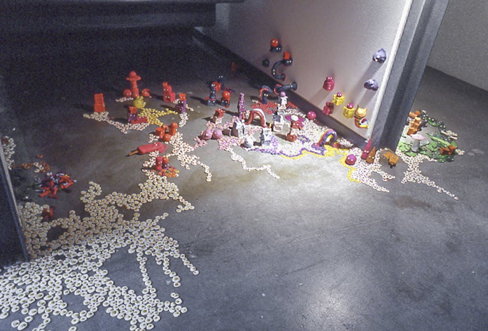 A series of small sculpture-like artworks are installed on a floor. A number of fried-egg shaped objects, red building-like figures, and small green round objects are placed widely on the floor.
