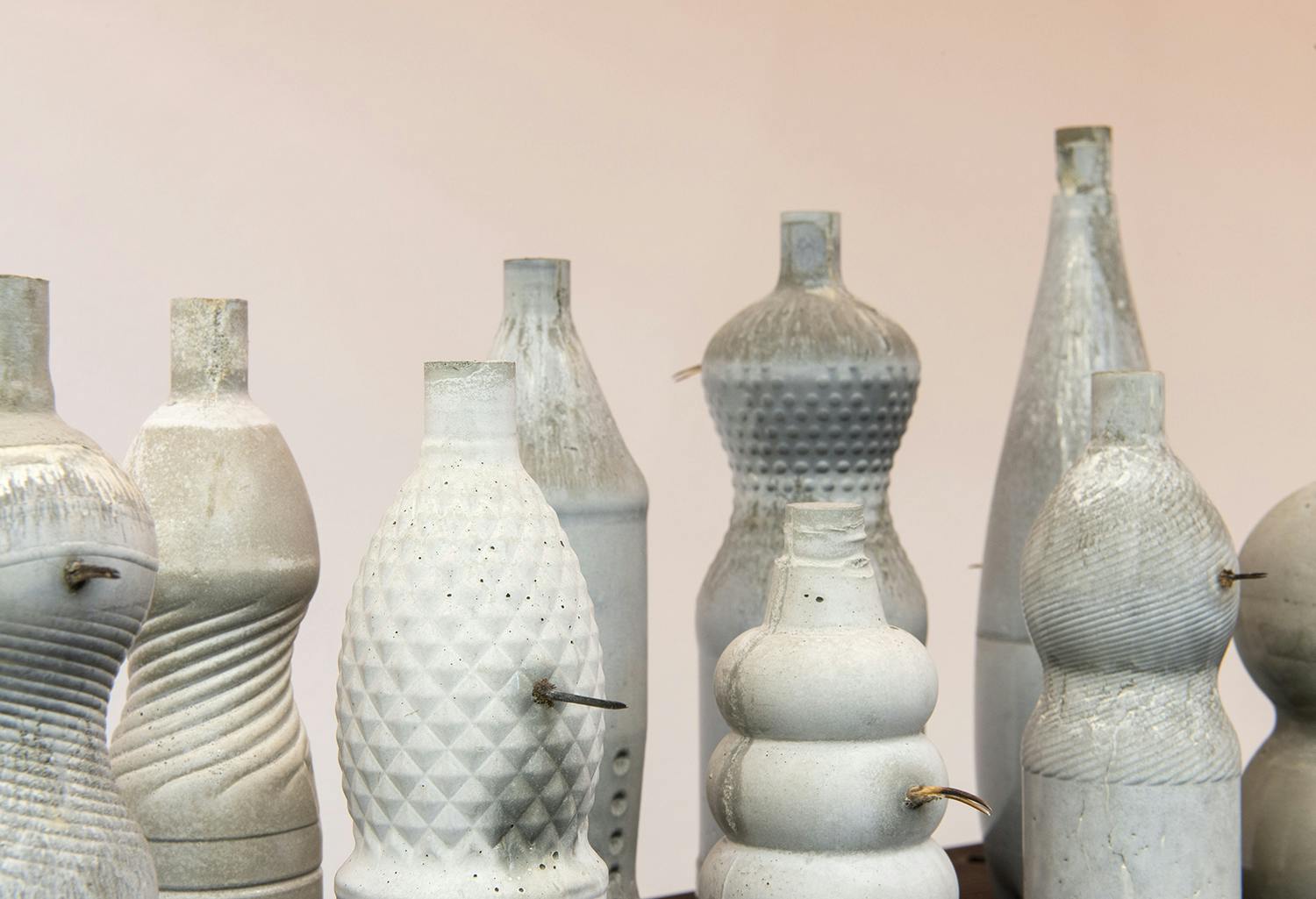A detail of an art installation by Corbin Union. White ceramic bottles are placed on a white wooden surface. A beak-like metal piece is sticking out of each ceramic pieces. 