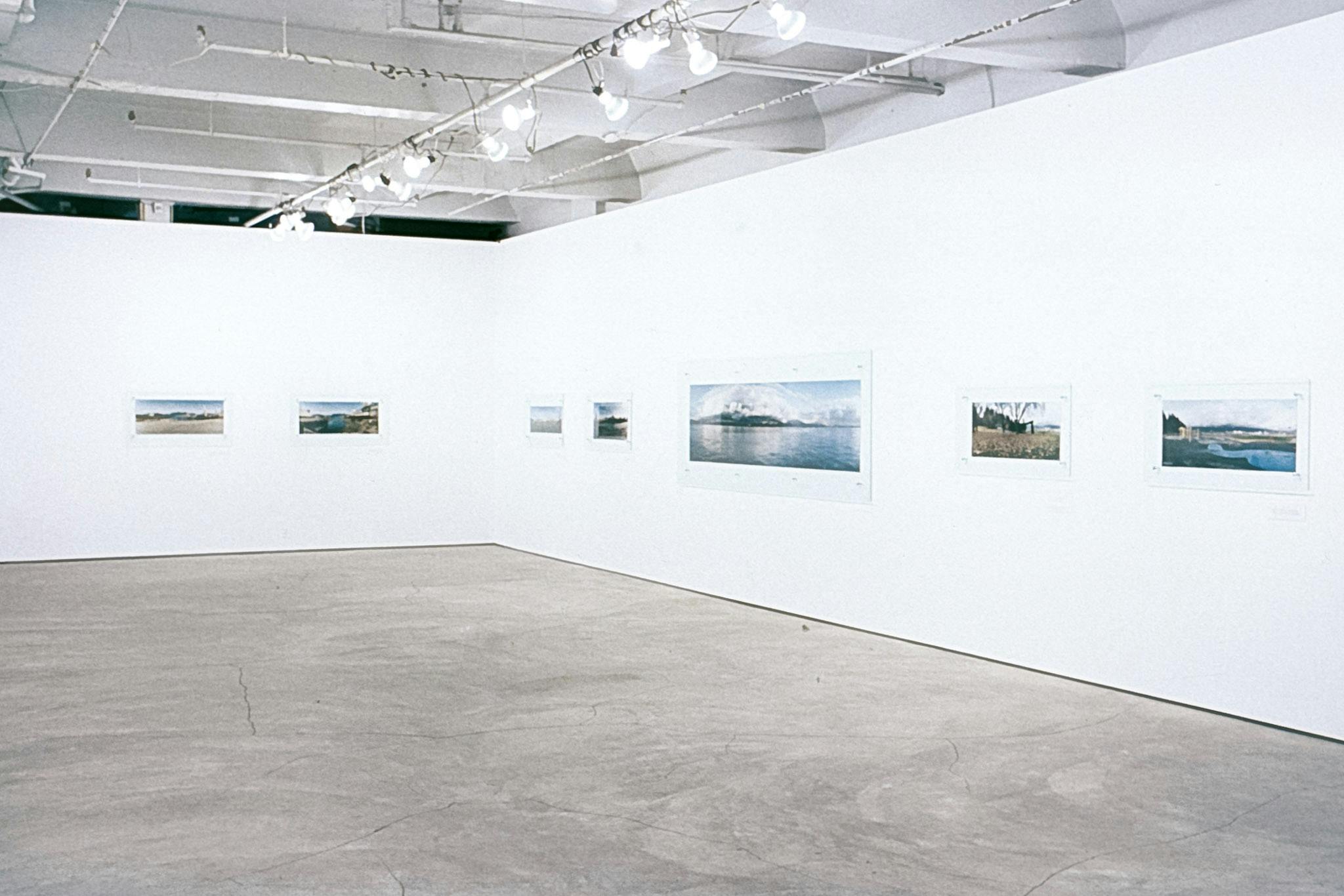 Several photos between sheets of glass mounted on 2 white walls. The photos vary in scale and show different landscapes across what is colonially known as the city of Vancouver. 