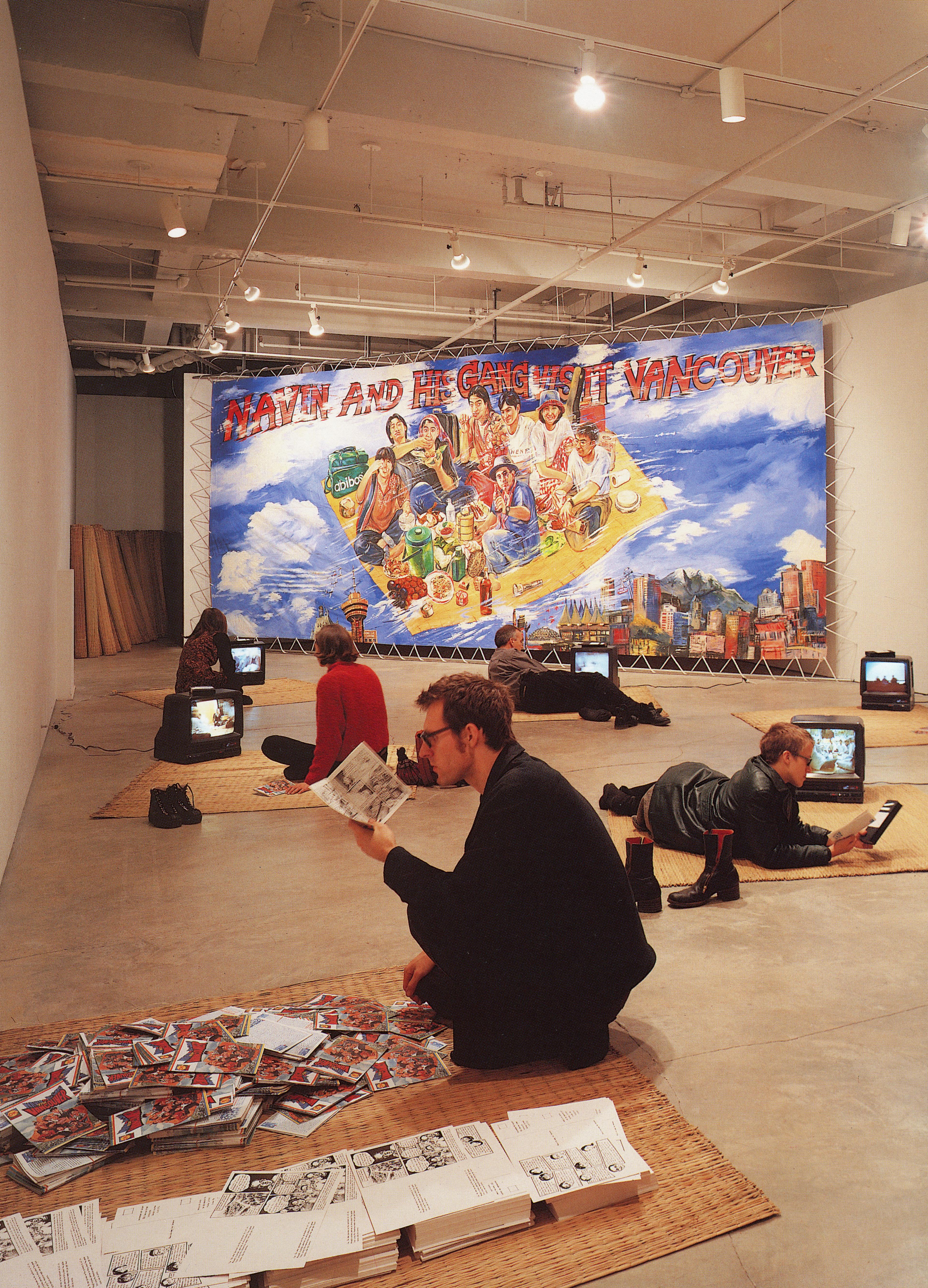 Several rush mats and CRT TVs are placed on the floor of a gallery space. Some visitors sit on the mats and watch videos. A large artist-made poster is mounted on the wall behind the installation. 