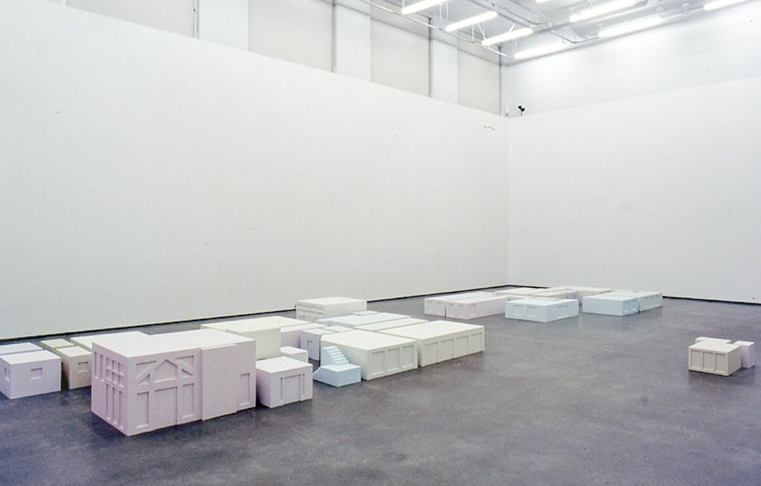 Many sculptures are installed on a gallery floor.  They are square shaped and low in height. Those cube-shaped sculptures are pale coloured; some of them are pale pink, yellow, blue, or green.  