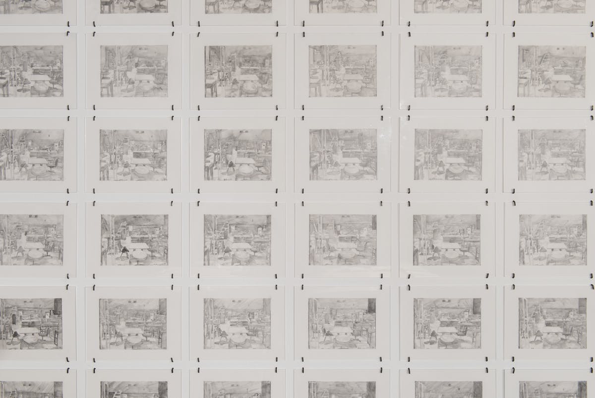 A close view of many small graphite drawings hung in a grid. Each drawing is the same depicting an interior view containing chairs, tables, ladders and other furniture.
