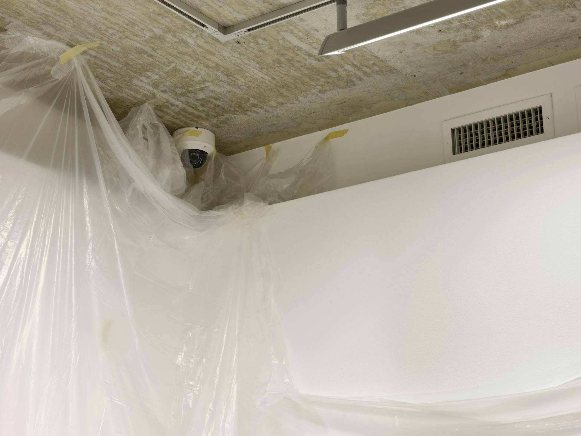 A security camera is surrounded by sheets of translucent plastic sheeting taped to the walls and ceiling in the corner of a gallery. 