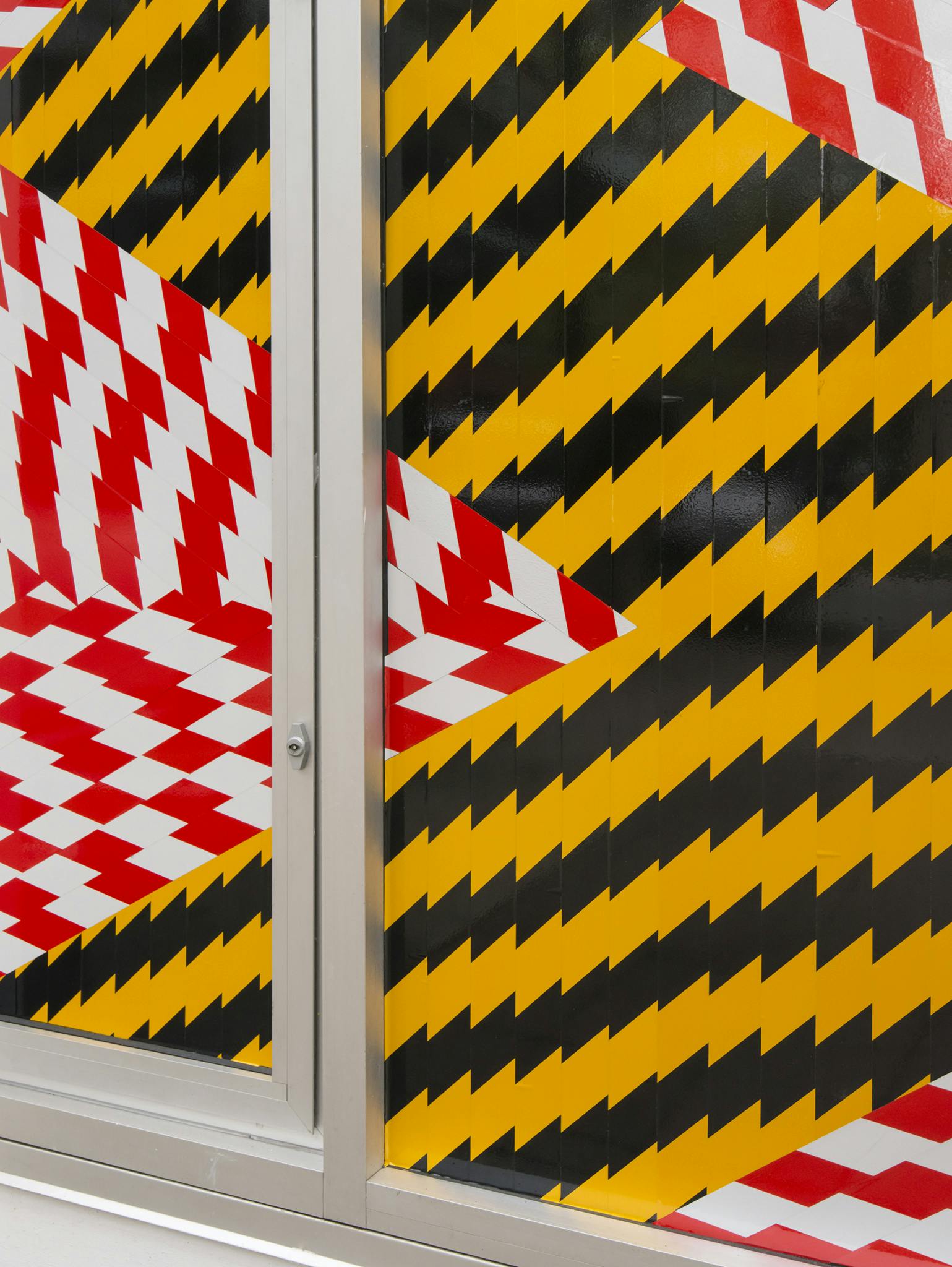 An image detail of red-and-white and black-and-yellow caution tape installed across CAG’s ground floor façade windows. The caution tape forms zig-zag lines and shapes. 