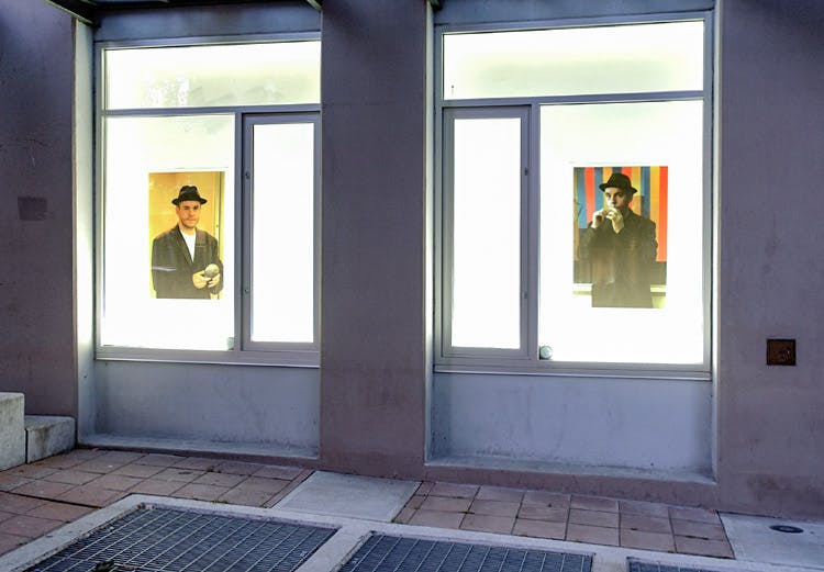 An installation image of a work by Daniel Olson in CAG’s window spaces facing a sidewalk. Two poster-shaped photography works are mounted on an illuminated wall behind glass windows. 