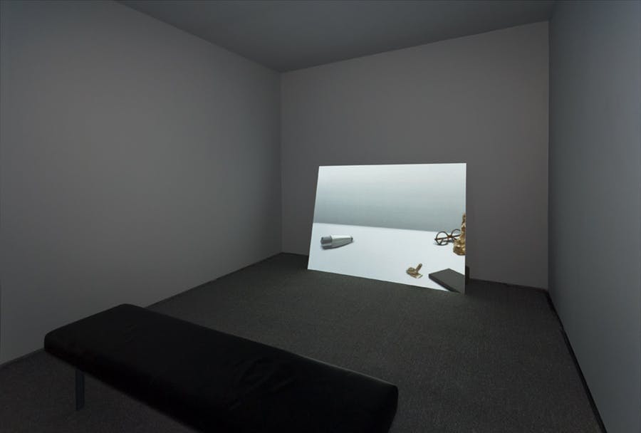 A bright, projected image appears to lean against the wall of a dimly lit gallery. The image displays several objects scattered on a floor. A black bench sits in the center of the room. 