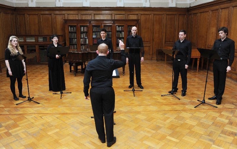Seven people in black outfit standing and performing a song inside a study. Six of them are the singers facing toward the conductor, who is raising their right arm. 