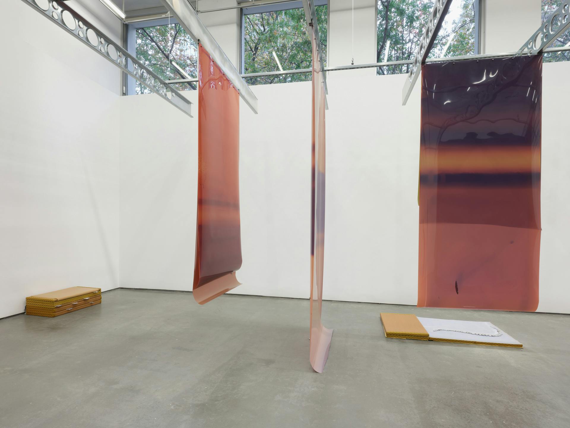 Long earth tone sheets of film of various colours are suspended from industrial metal ceiling beams. Two sculptures, made of tatami mats and cast aluminium objects, sit on the cement floor in the background. Large windows are visible near the ceiling.