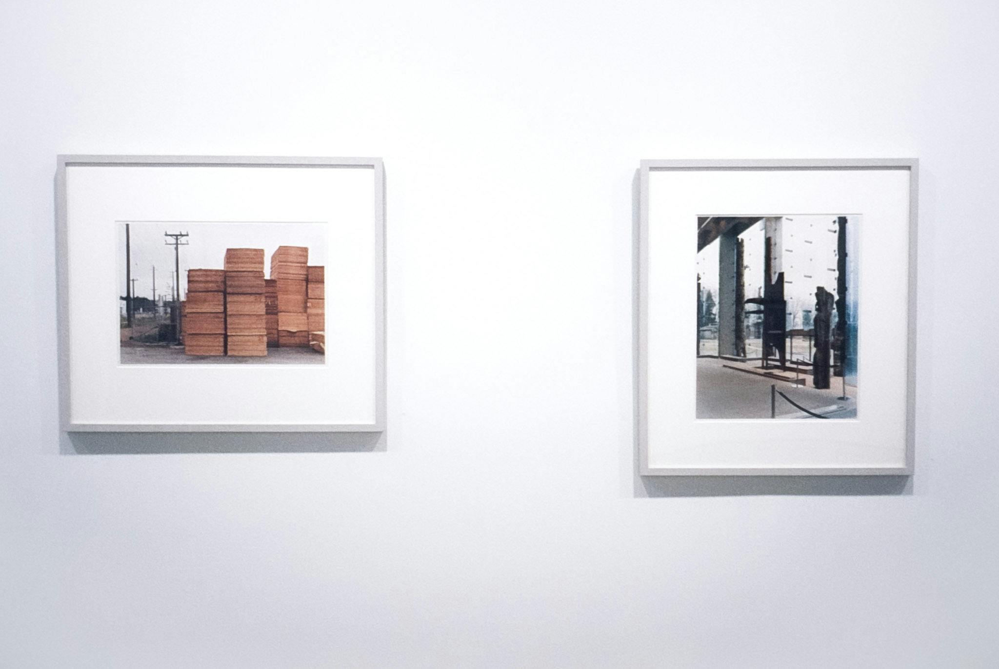 A closeup of 2 framed photographic works on a white wall. On the left, a horizontal photo shows stacks of plywood and cell towers. The vertical one on the right, shows what looks like a museum lobby.