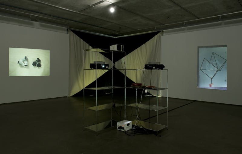 Two glass shelves installed in the middle of a gallery hold five slide projectors. Two images are projected onto the gallery walls. A rectangular fabric piece hangs in the corner between the images.
