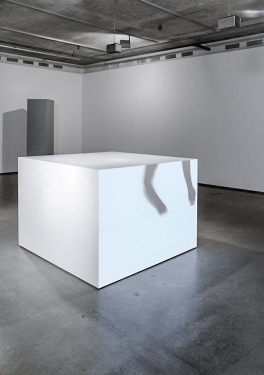 A large white cube is installed in the middle of a gallery space. A shadow of two long narrow objects hanging from the ceiling cast on one side of the cube.