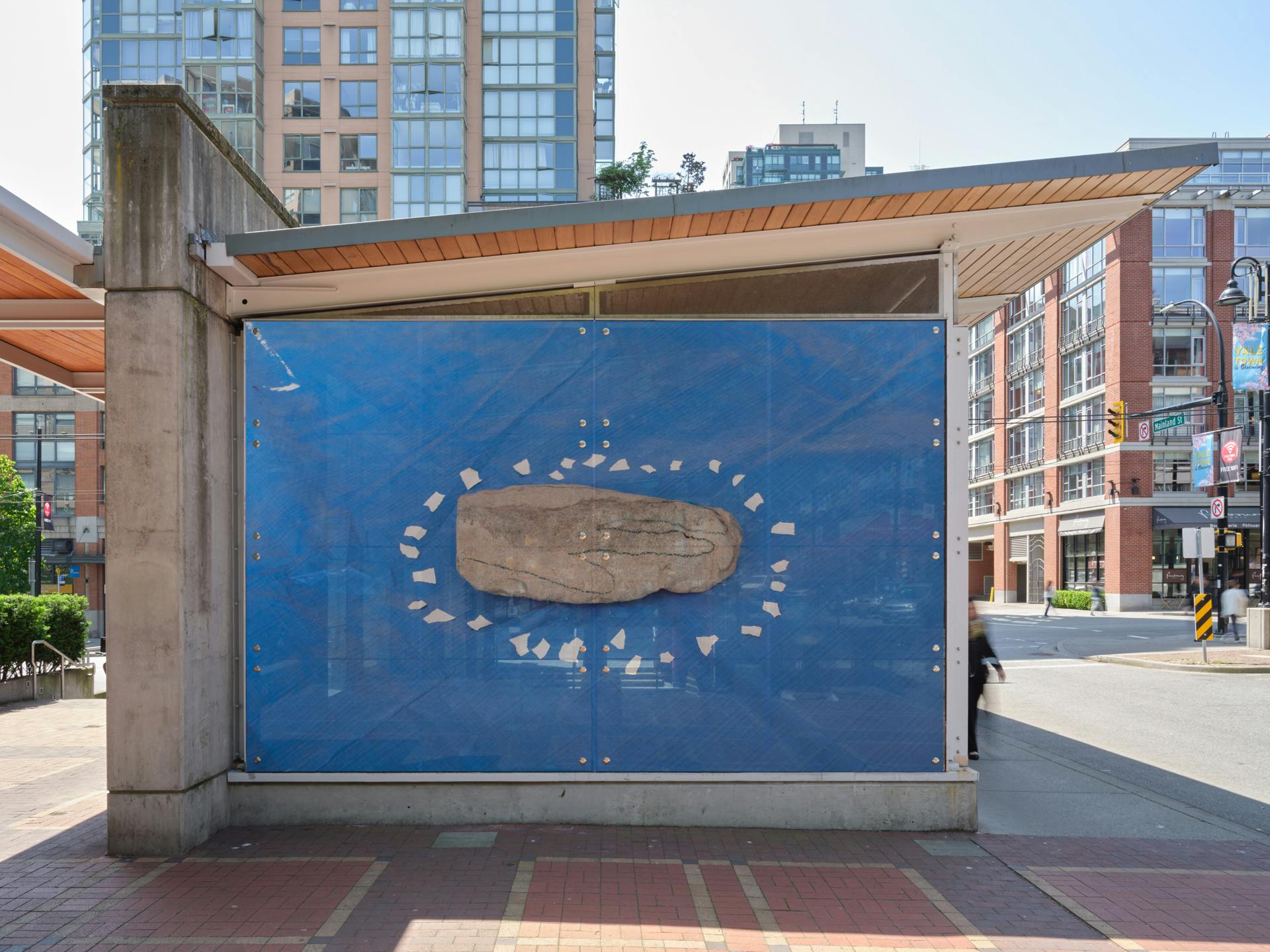 A frontal view of a SkyTrain station with one photo in its window. The photo depicts a rock on a blue tarp with a hand drawn on its surface and bits of white tape arranged in an oval around its perimeter. 