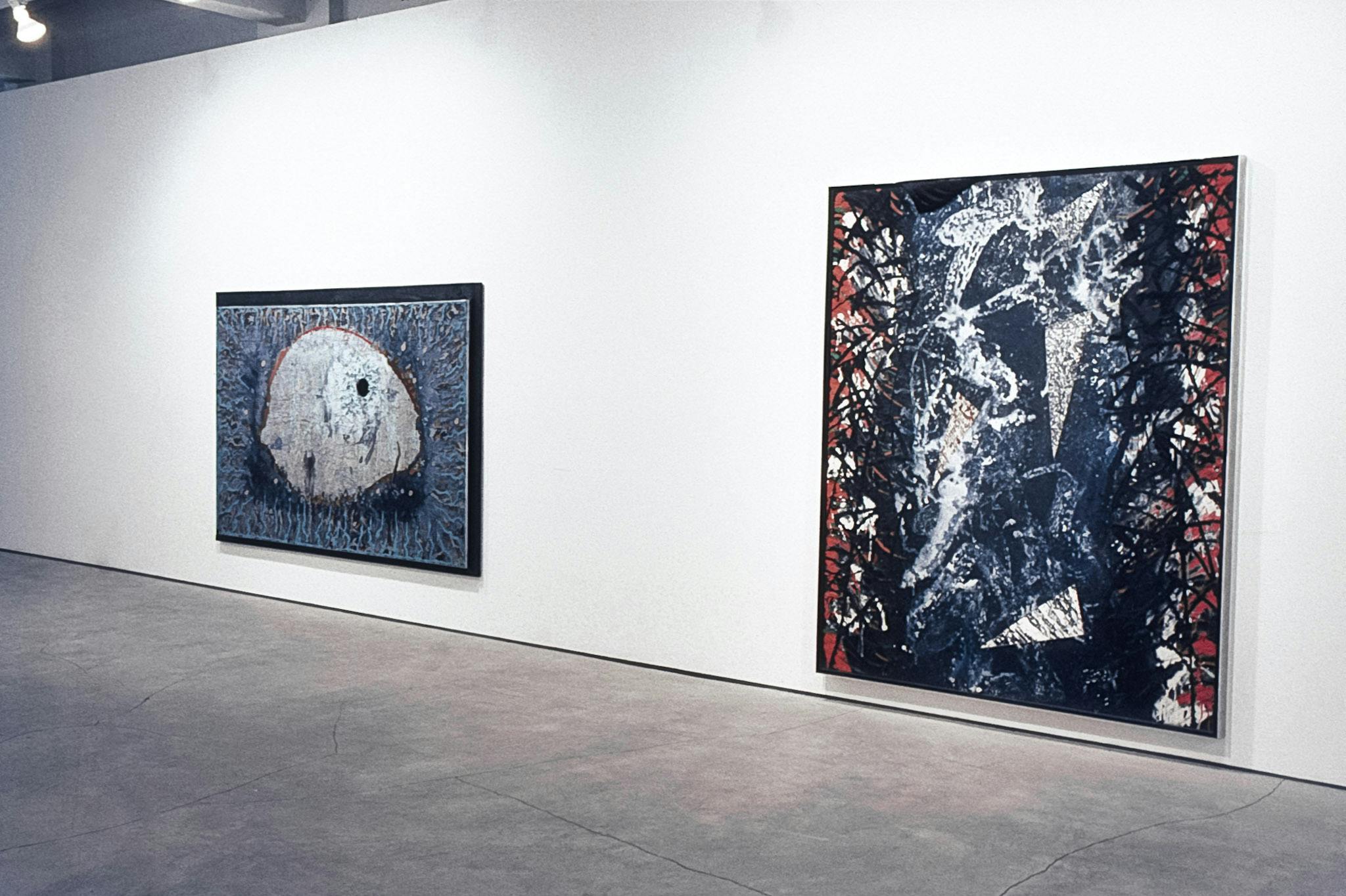 2 paintings on a wall. The one in the foreground has red edges and a black centre. The surface is made by splattering and swirling paint. Small, triangular strips of material can be seen throughout. 