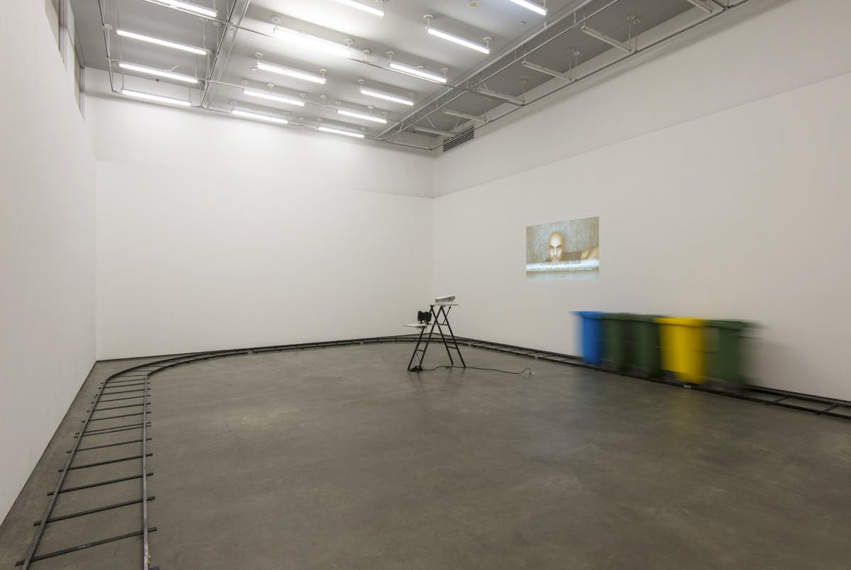 Image of a gallery space. A video is projected on a wall. A small rail line installed around the perimeter of the gallery, on which five connected garbage bins of various colours move. 