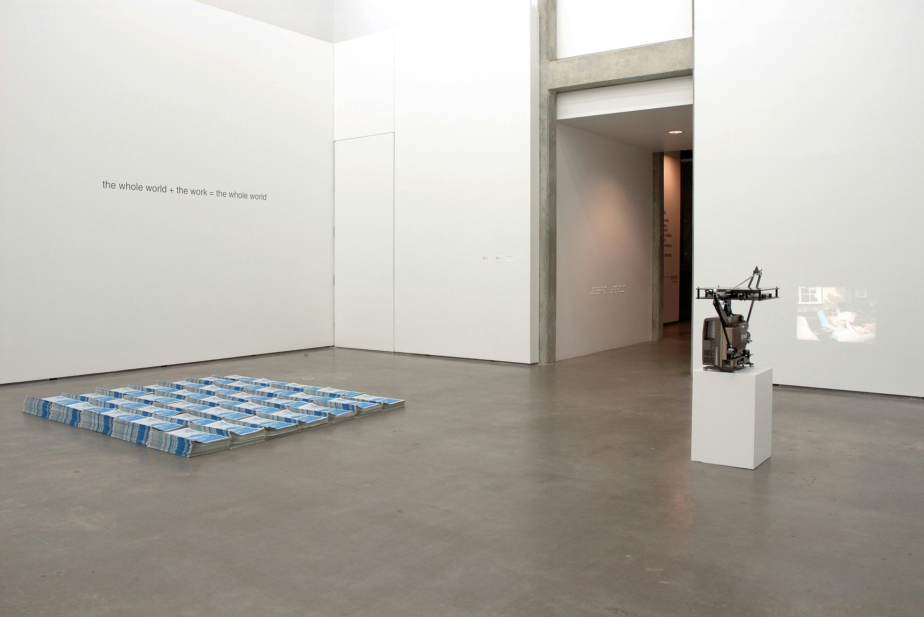 This is an installation shot of a group exhibition titled Concrete Language. Stacks of brochures are placed on the gallery floor. On a wall behind brochures, a text-based artwork is printed. 