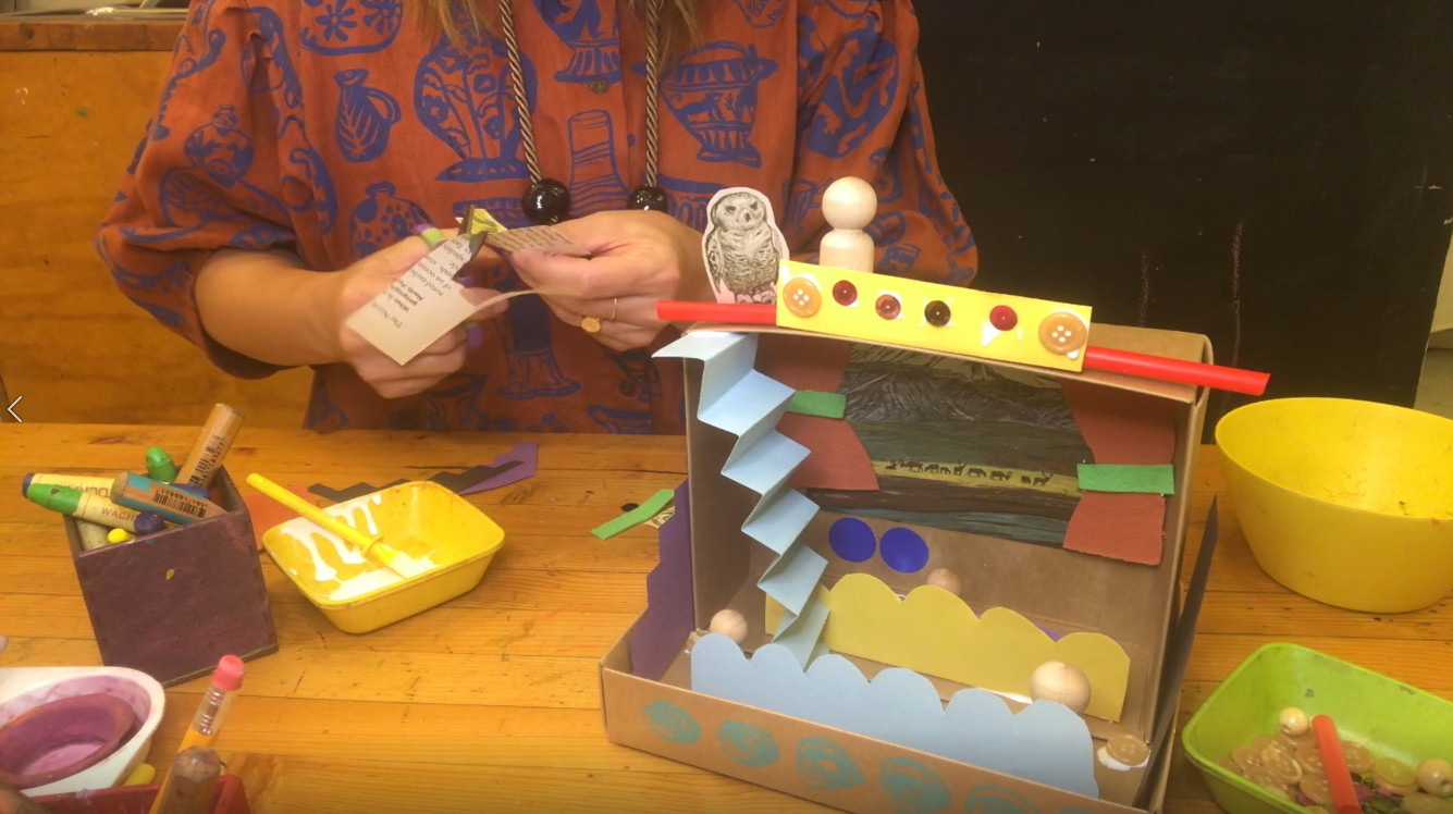A still of a person making a theatre set with cardboard boxes,  colourful construction paper, and wood beads. 
