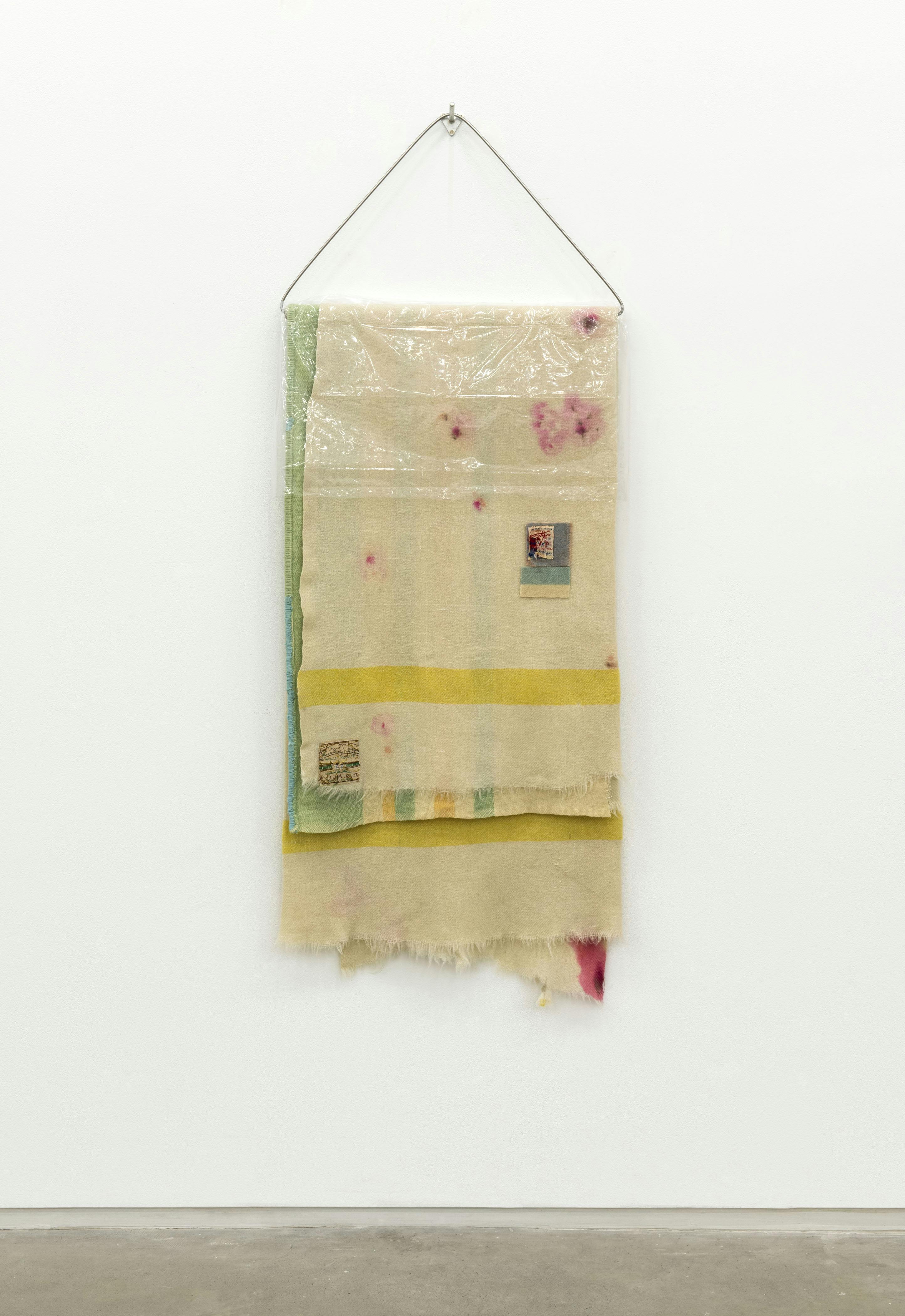 A cream coloured textile with yellow and green stripes hangs from a triangular metal armature mounted on a gallery wall. Pink splotches and small patches of textiles are scattered around the surface. 