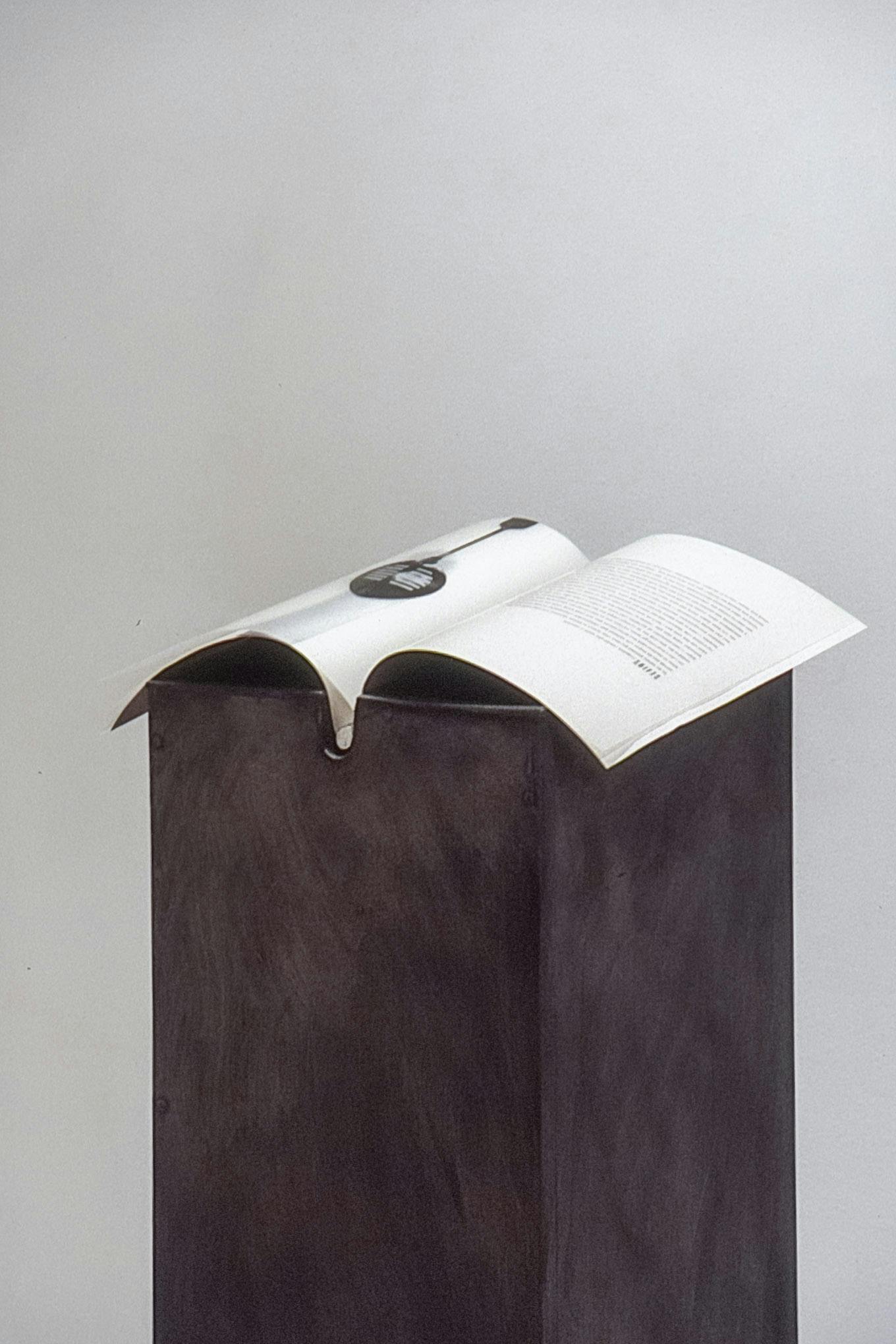 A thin, open book rests on the opening of a dark, brushed metal plinth. The plinth appears to be hollow and has a small divot to hold the spine of the book. The background shows a white wall.