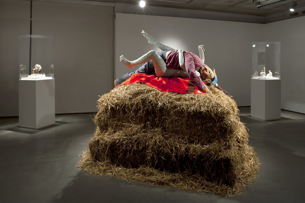 A large pile of straw sits on a gallery floor on which two, embracing mannequin-like forms are laid. Two small sculptures, each under glass atop a plinth, are visible in back of the exhibition space.