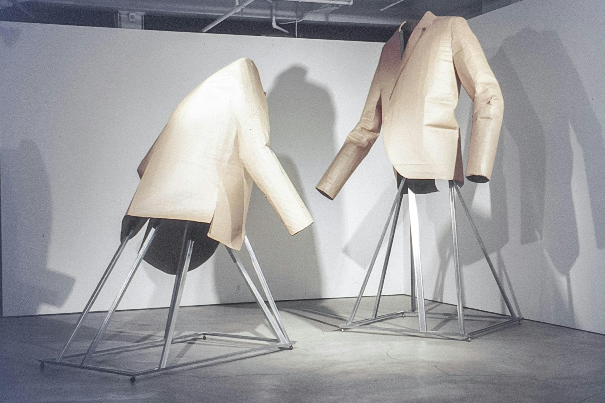 Two large sculptures in a gallery. The works are made of metal pyramidal frame bases, with huge blazers made of cardboard at the top. The blazers face each other and one is leaning forward slightly. 