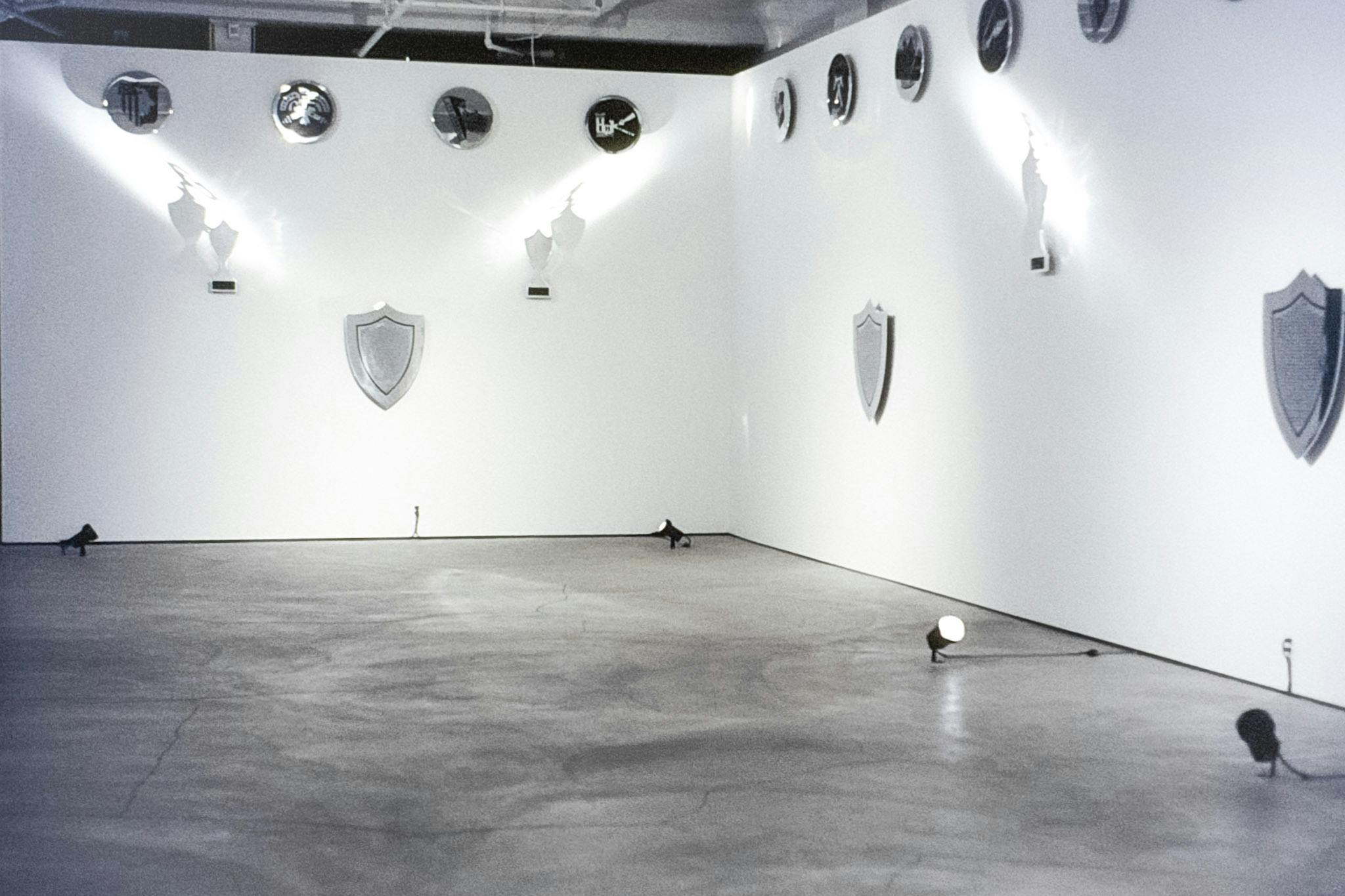 The corner of a gallery space with several metal artworks mounted on the walls. At the top there is a row of circular crests and below there are shield and trophy-like shapes. 