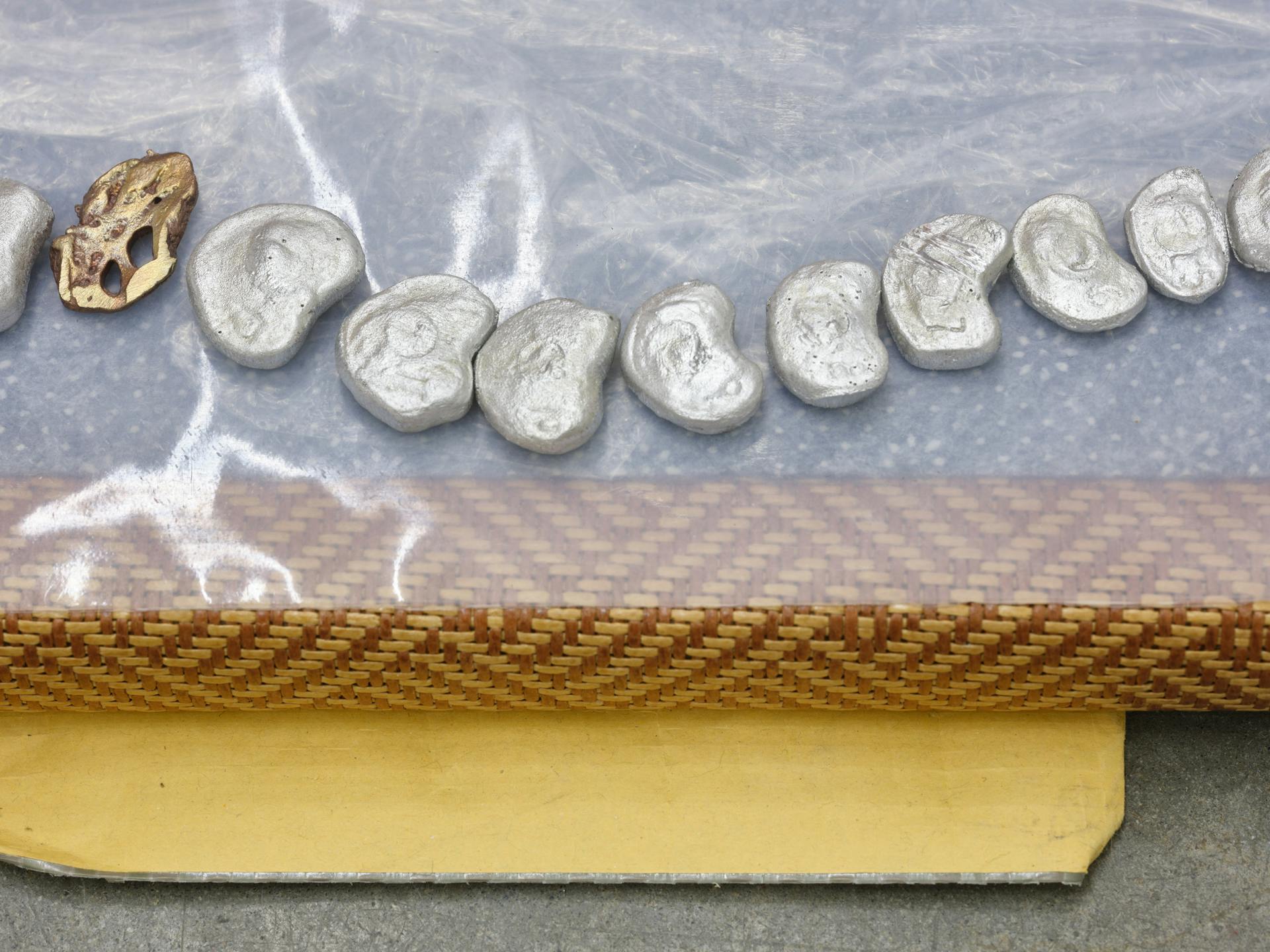 Detail view of a patterned tatami mat with one bronze and ten silver discs arranged in a curved line on its surface. The edge of a manila envelope is tucked underneath the mat.