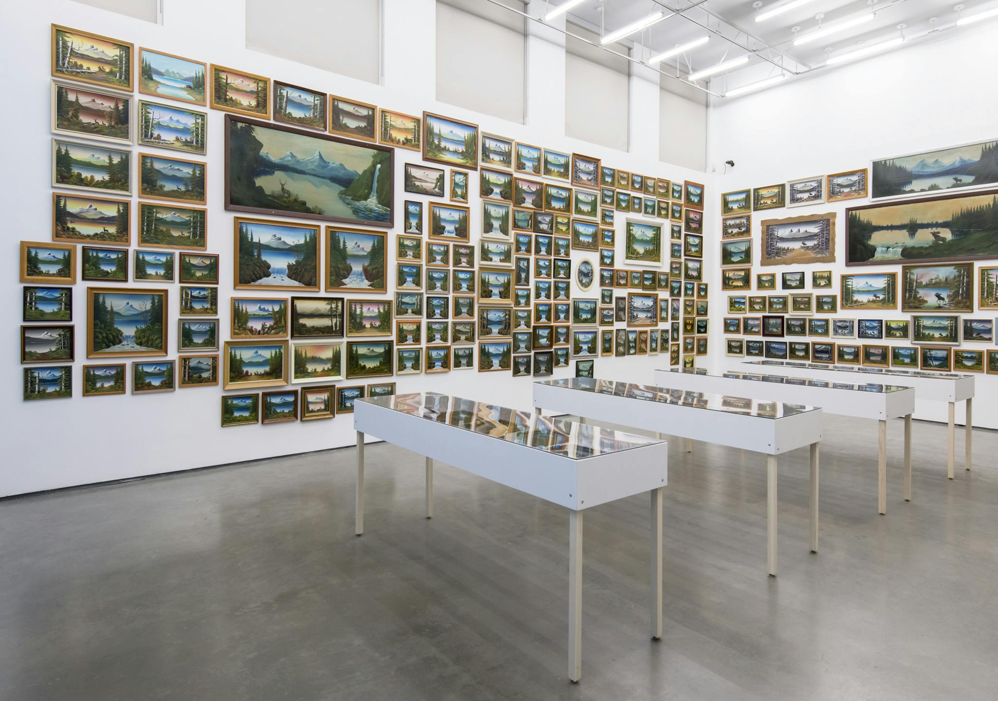Multiple, framed paintings of similar mountain landscapes in different sizes cover the walls of a gallery in a salon-style layout. Four vitrines in the center of the gallery display more. 