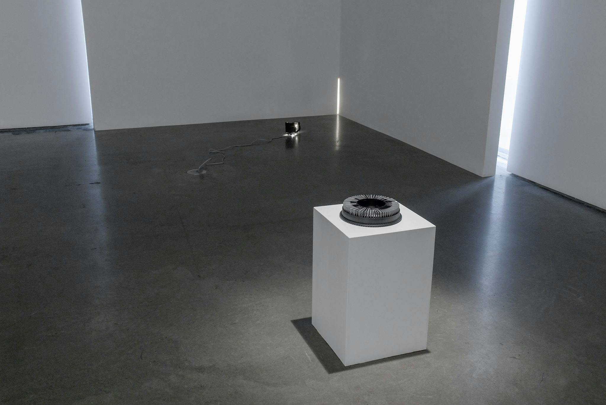 A black carousel slide tray is placed on a pedestal installed in the middle of a gallery space. A tiny light is also installed on the floor around the corner of the same room. 