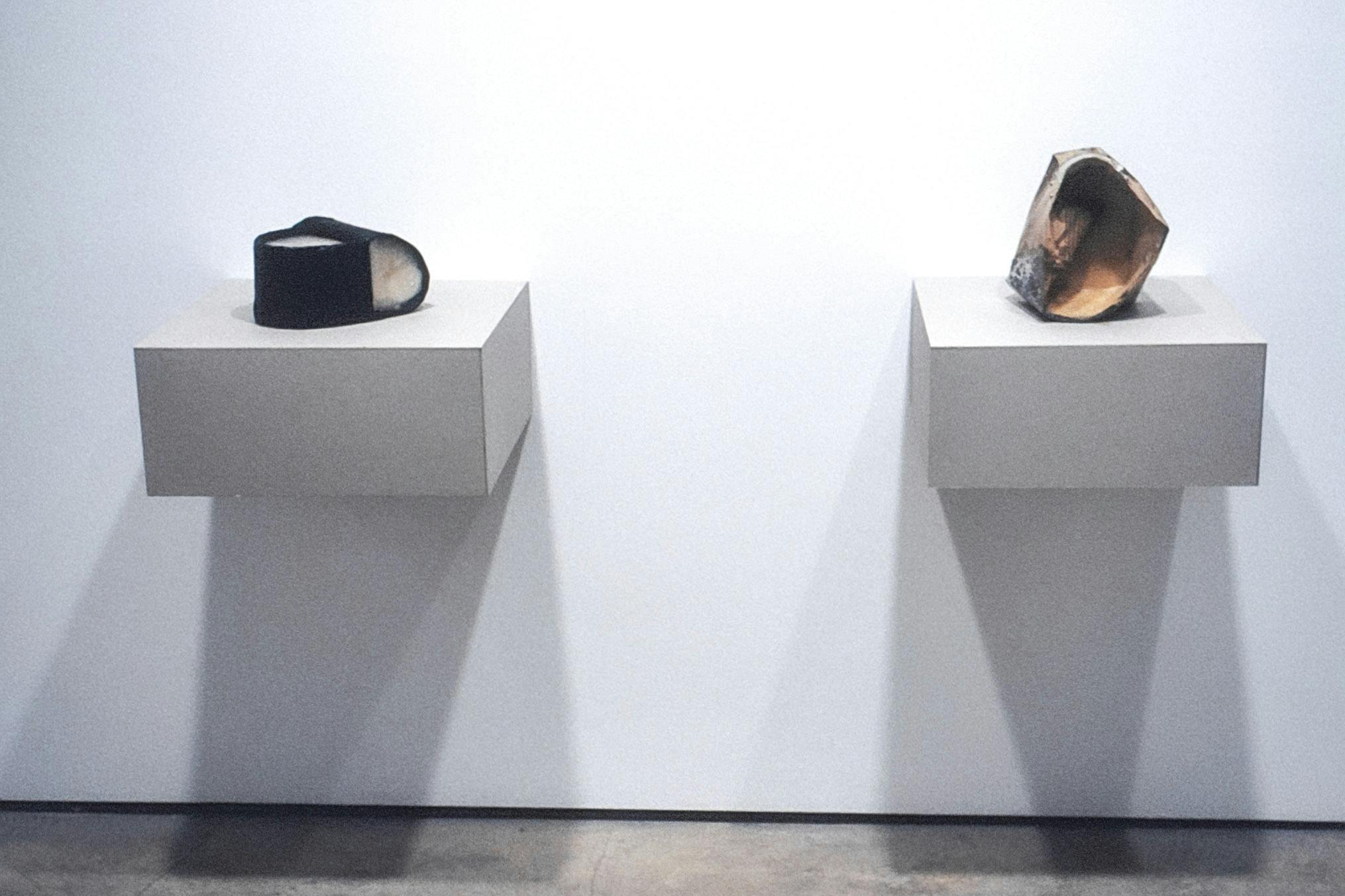 2 artworks on small grey plinths mounted on white walls. On the left is a black vessel with white on its "handle" and white filling inside. The other is a carved piece of wood with a concave interior.