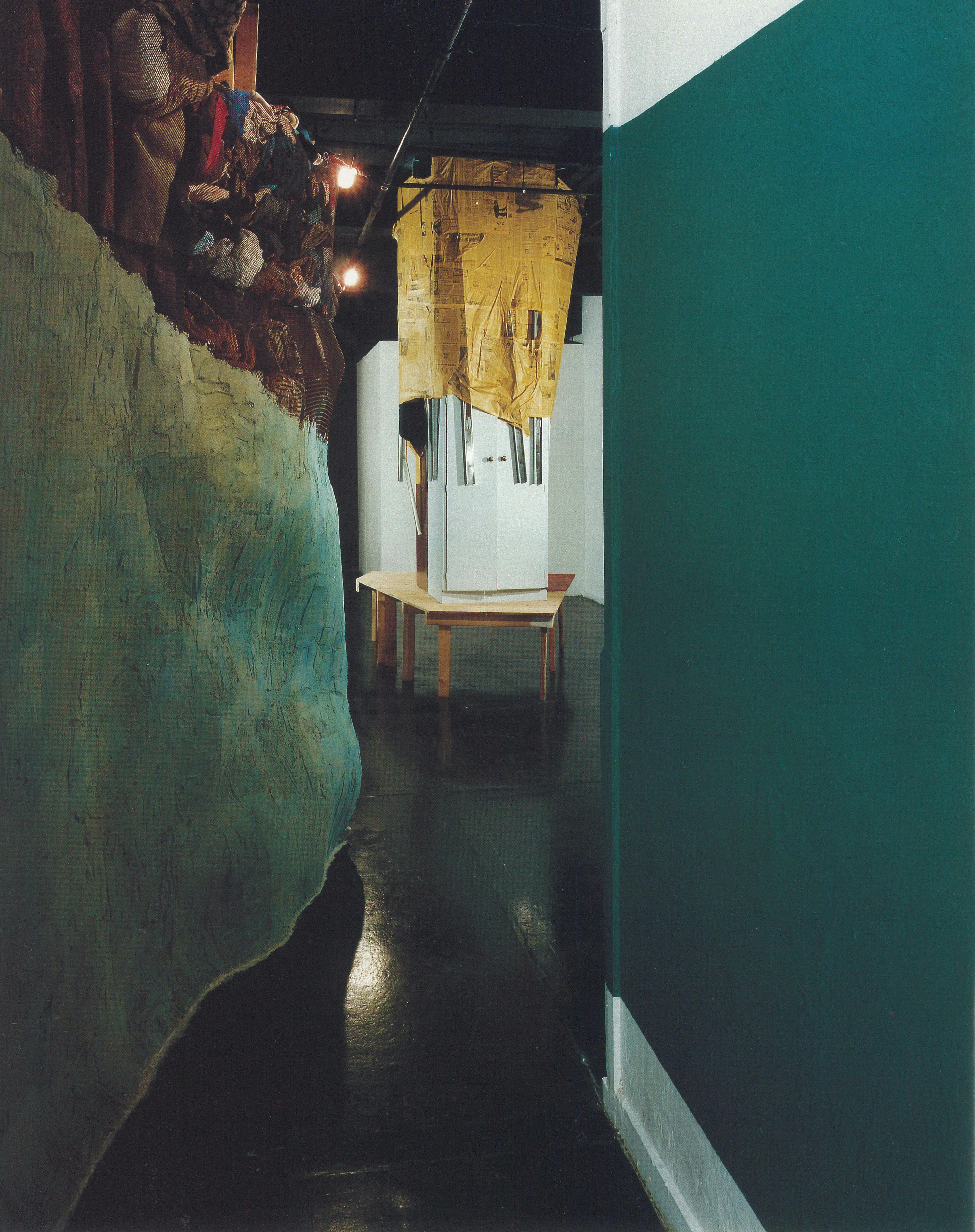 In a gallery space, two green painted walls lead to a space where a pentagonal-shaped wooden table sits. A white closet is placed on top of the table. The closet is half-covered by old newspapers.