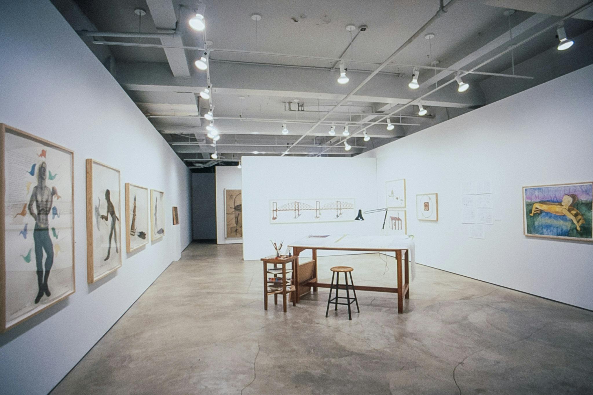 Various artworks are installed in the galley. Five drawings of human figures are mounted on the walls. A large wooden desk with a stool is installed in the middle of  the floor. 