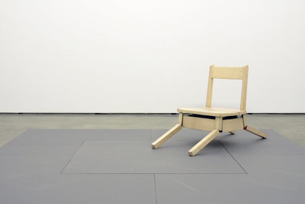 A small mechanized wooden chair is installed on the gallery floor. This photograph captures the moment in which the chair is about to rise up from the posture of a dog lying down.