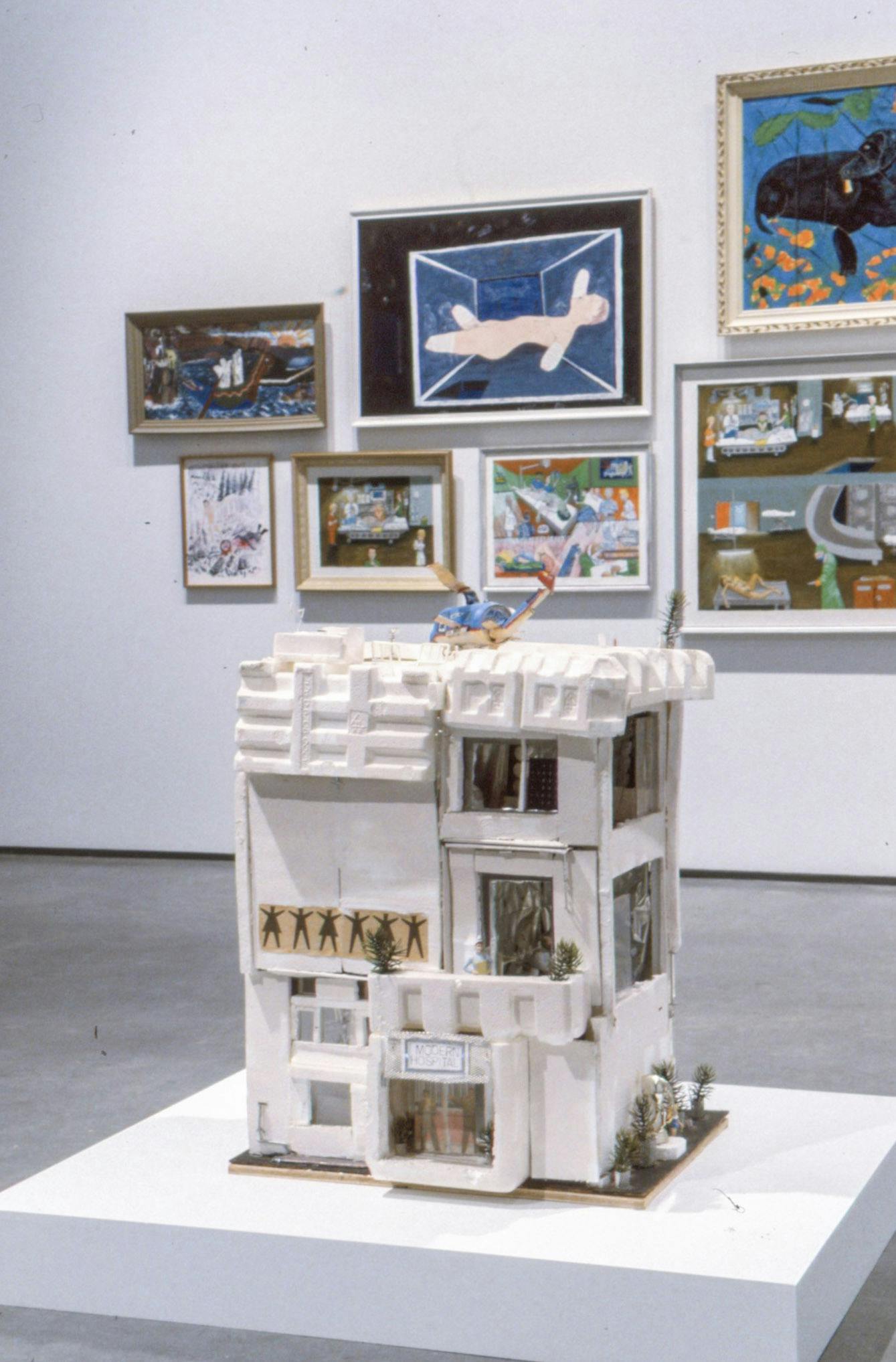 A series of framed paintings are mounted on the gallery wall behind the sculpture. The sculpture depicts a white apartment building, on which a blue helicopter is landing. 