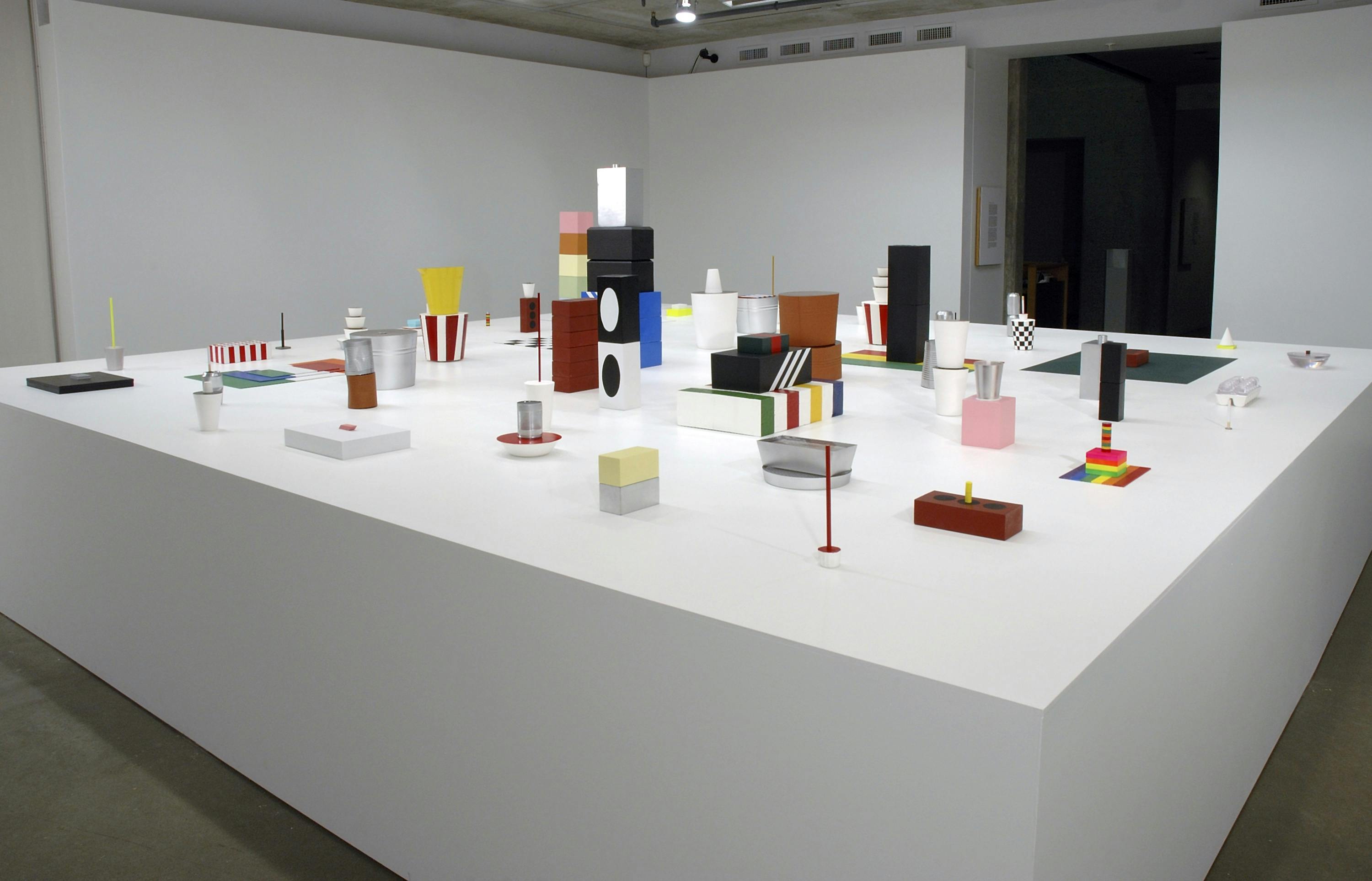 Install image of sculptures on a large low pedestal placed in a gallery. Sculptures vary in size, shape, and colour. They mimic commercial objects and icons such as a Kentucky Fried Chicken bucket and Hudson’s Bay stripes.