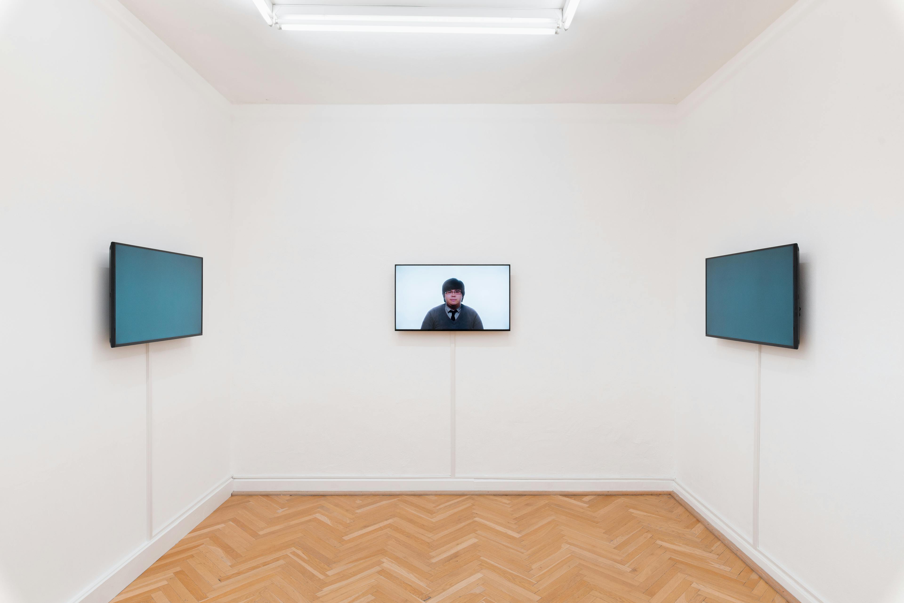 A small gallery room with three monitors on three walls. On the centre back wall, the monitor plays a video of a talking head. On the parallel walls, two monitors with blue screens face each other.
