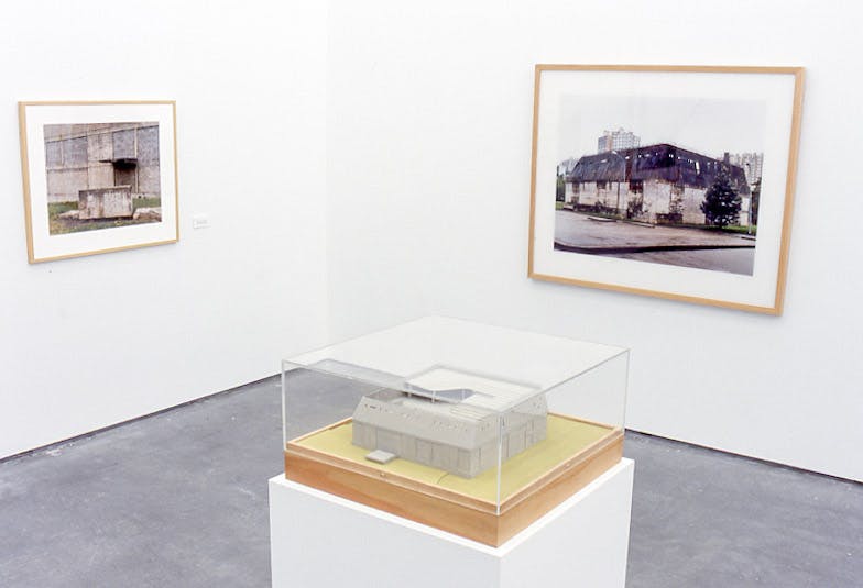 A closeup of a corner in a gallery. There are 2 framed pictures showing run-down buildings, and a plinth with a glass case. The plinth holds a miniature model of a building in one of the photos.