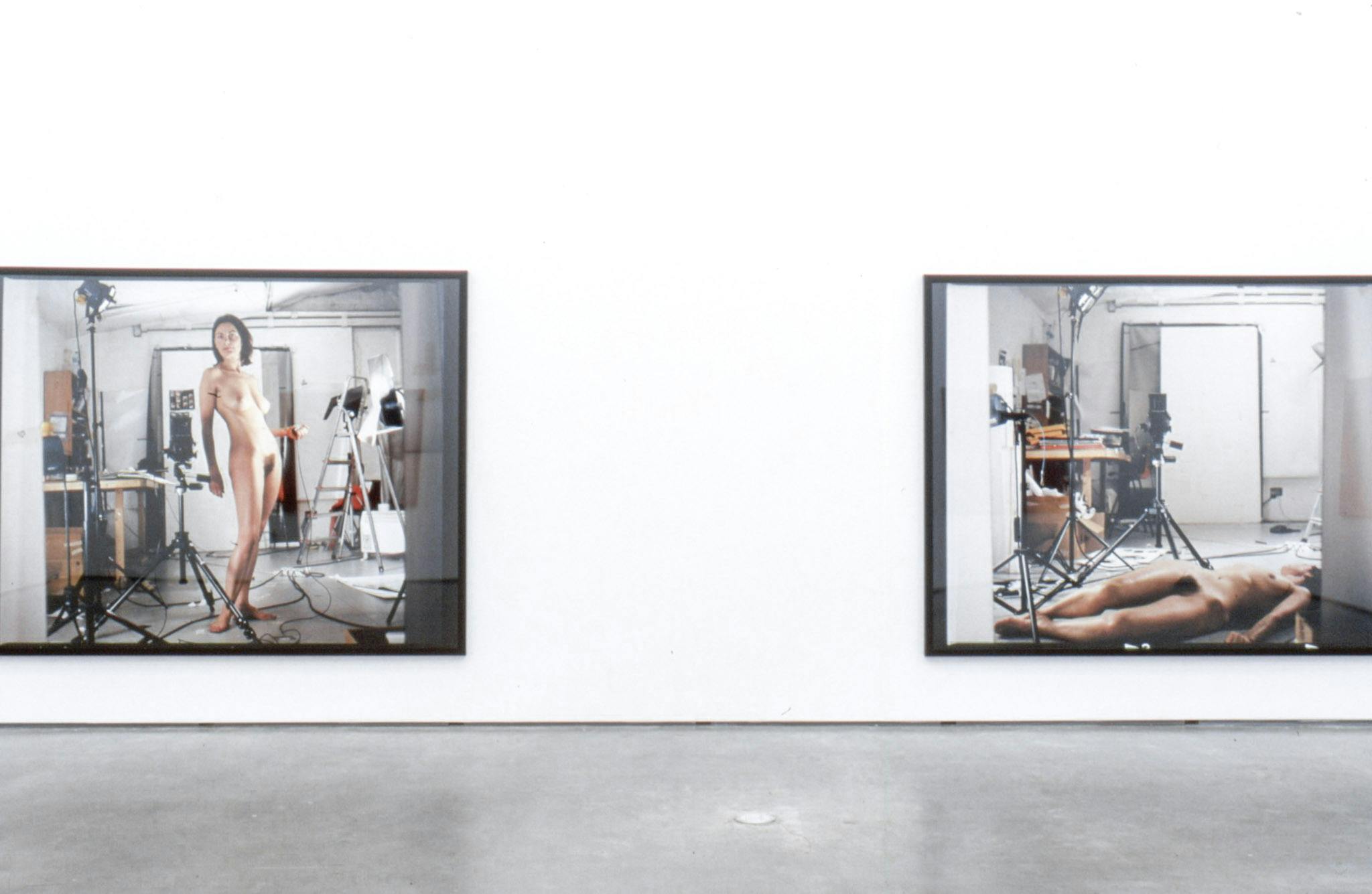 Two large-sized photographs are installed on a gallery wall. Both photographs show a naked woman posing in the middle of an artist studio. One image shows the person lying down on the floor.