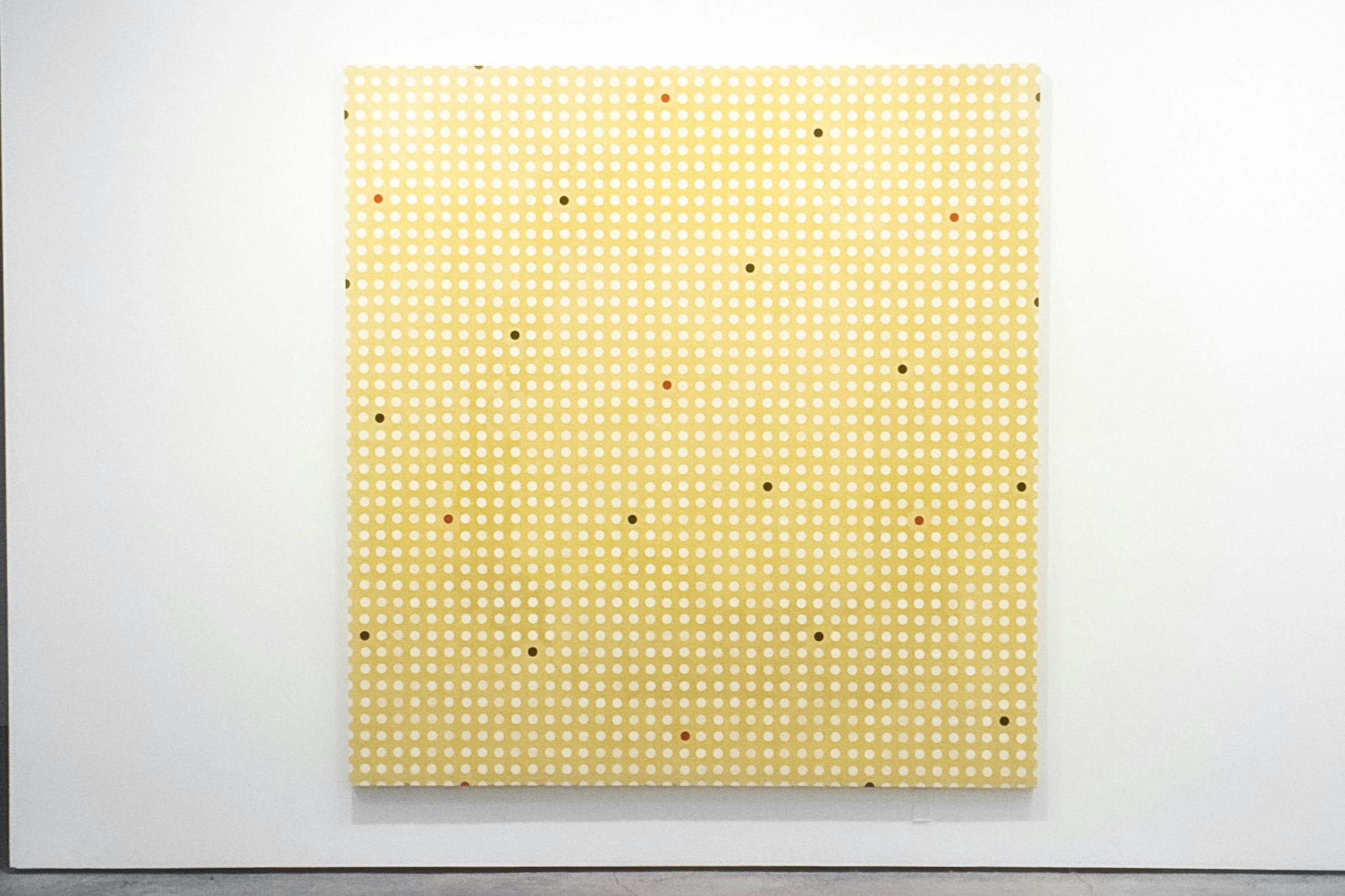 A large painting mounted on a white wall. The painting has a yellow background and a gridded dot pattern. Most of the dots are white, but some red and black dots are spread throughout.