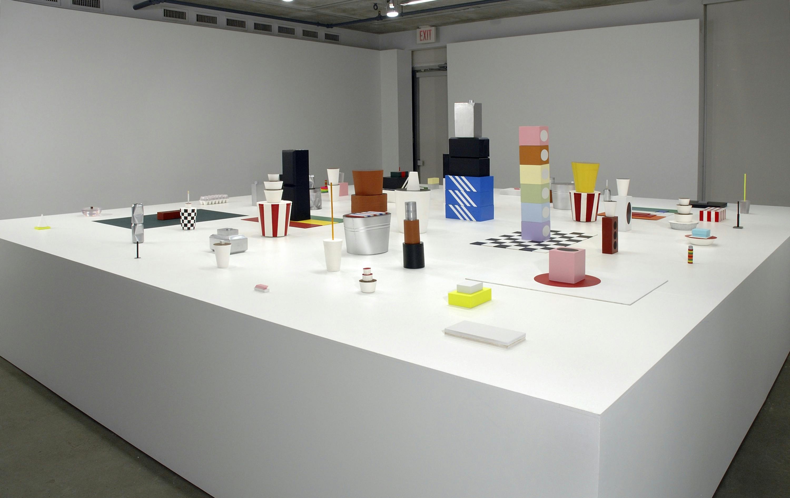 Install image of sculptures on a large low pedestal placed in a gallery. Sculptures vary in size, shape, and colour. They mimic commercial objects and icons such as Kentucky Fried Chicken bucket and Hudson’s Bay stripes.
