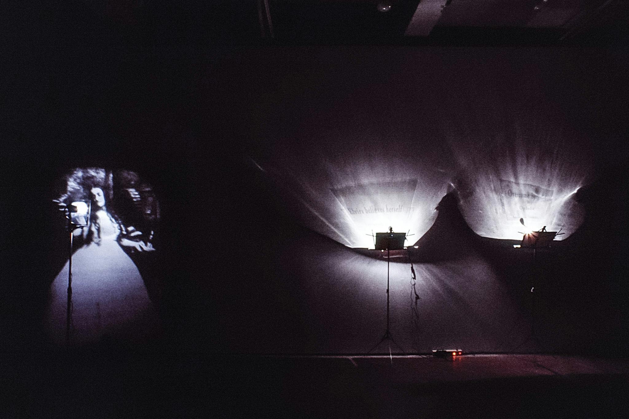 An installation image of a dimly-lit gallery space. A pair of stage lights are installed closely to each other near the wall. An image of a person in a dress is projected next to that pair of the lights. 
